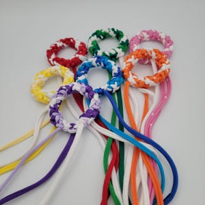 May include: A collection of colorful, braided fabric rings with attached cords. The rings are in various colors, including red, green, pink, orange, yellow, purple, and blue, each with a white accent. The cords are also in matching colors.