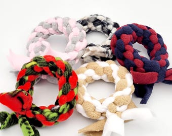 May include: Five braided dog toys in different colors and patterns. The toys are shaped like wreaths and have a knotted bow on one end. The colors include pink, black and white, red and blue, green and red, and tan and white.