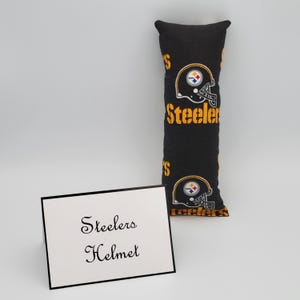 May include: Black and gold Steelers helmet pillow with the team logo and the word "Steelers" repeated on the fabric. The pillow is sitting on a white surface with a white card that says "Steelers Helmet".
