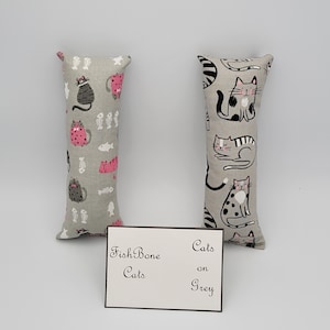 May include: Two gray cat-themed catnip toys. One toy has a pink and black cat pattern with fish bones and fish. The other toy has a black and white cat pattern. The text on the card reads "Fish Bone Cats on Grey".