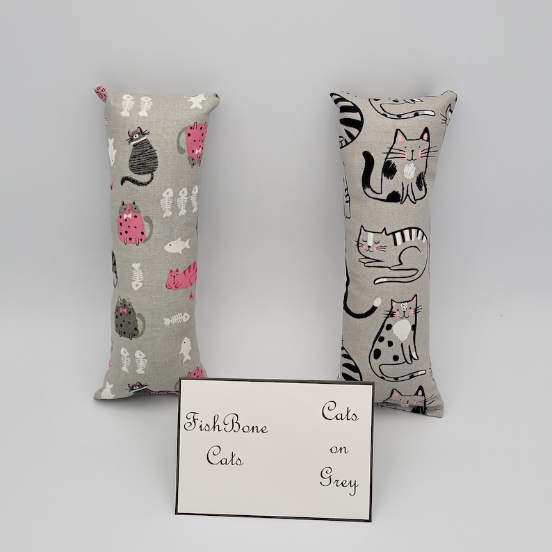 May include: Two gray cat-themed catnip toys. One toy has a pink and black cat pattern with fish bones and fish. The other toy has a black and white cat pattern. The text on the card reads "Fish Bone Cats on Grey".