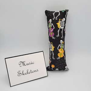 May include: A black and white pillow with a repeating pattern of skeletons playing musical instruments. The instruments include a saxophone, a cello, and a bass. The text "Music Skeletons" is printed on a white card in front of the pillow.