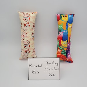 May include: Two cat-themed fabric pillows. The pillow on the left has a cream background with a repeating pattern of red and white oriental cats. The pillow on the right has a red background with a repeating pattern of colorful smiling cats. The pillows are labeled "Oriental Cats" and "Smiling Rainbow Cats".