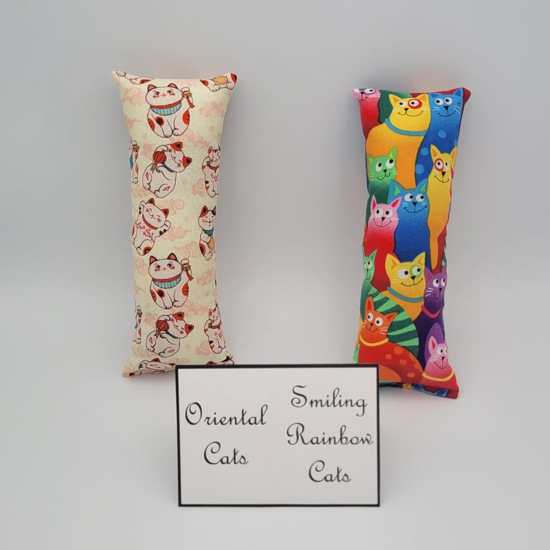 May include: Two cat-themed fabric pillows. The pillow on the left has a cream background with a repeating pattern of red and white oriental cats. The pillow on the right has a red background with a repeating pattern of colorful smiling cats. The pillows are labeled "Oriental Cats" and "Smiling Rainbow Cats".