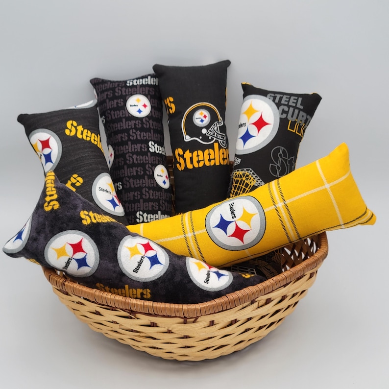 May include: A wicker basket filled with six decorative pillows featuring the Pittsburgh Steelers logo and team colors. The pillows are made of black and yellow fabric with a variety of patterns, including a plaid design and a repeating Steelers logo.