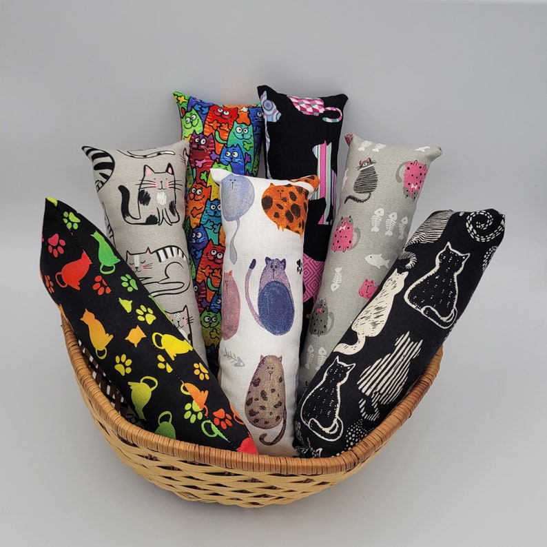 May include: A collection of eight cat-themed catnip toys in a wicker basket. The toys are made of fabric and feature various cat designs, including a black cat, a white cat, and a colorful cat. The toys are all different shapes and sizes.