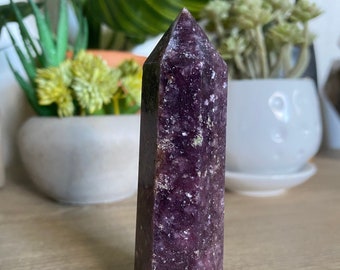 May include: A tall, purple lepidolite crystal tower with a pointed top. The crystal has a rectangular shape with visible texture and small white and green inclusions. The crystal is standing on a light brown wooden surface.