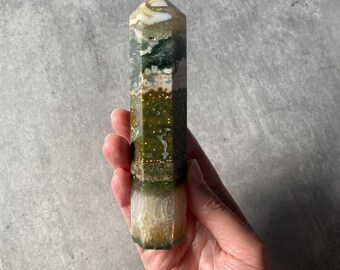 Puede incluir: Un obelisco de piedra preciosa pulida con forma hexagonal. La piedra presenta una mezcla de colores, incluyendo verde, blanco, beige y marrón. El obelisco se sostiene en una mano sobre un fondo gris.