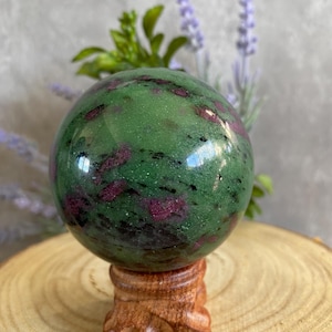 Könnte beinhalten: Eine grüne und rosafarbene Zoisit-Kugel auf einem Holzständer. Die Kugel ist poliert und hat eine glatte Oberfläche.