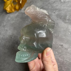 May include: A green and purple fluorite skull with a carved lizard on top. The skull is polished and has a smooth surface.
