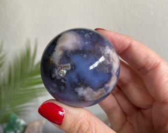 May include: A spherical gemstone with a marbled pattern of blue, white, and brown. The orb is held in a hand with red nail polish, highlighting the stone's reflective surface and natural colours. The gemstone is a decorative item.