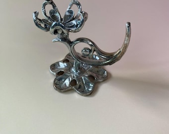 May include: A silver-toned metal wall hook featuring a decorative base with five circular cutouts and a curved arm that terminates in a stylised flower-shaped holder. The metallic finish has a slightly weathered, textured appearance throughout.