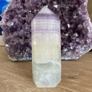 Peut inclure: Une pointe de cristal de fluorine vert clair et violet avec un sommet pointu et une base plate. Le cristal a un aspect stratifié avec des bandes de vert clair et de violet.