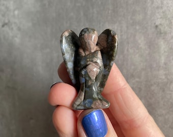 May include: A small, carved stone figurine in the shape of an angel with folded wings. The stone features a mottled pattern with earthy tones of dark gray, brown, and muted pink, displaying a smooth, polished surface and distinct natural mineral inclusions.