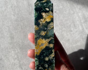 Puede incluir: Una torre de cristal rectangular pulida con una parte superior puntiaguda. La piedra presenta una mezcla de patrones verde oscuro, amarillo y blanco. El cristal se sostiene en una mano sobre un fondo neutro.