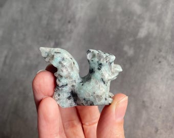 May include: A small, carved squirrel figurine. The sculpture is a light teal colour with black speckles, and is held in a person's hand. The squirrel is in a sitting position with its tail curved upwards. The background is a solid grey.