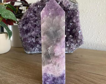 May include: A purple amethyst crystal point with a pointed top and a flat base. The crystal has a light purple colour with darker purple streaks throughout.
