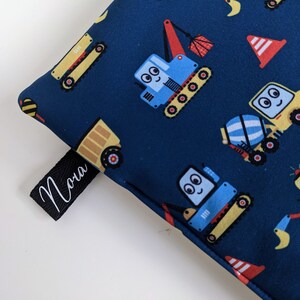 Könnte beinhalten: Eine blaue Stofftasche mit einem Druck von Baufahrzeugen. Die Tasche hat ein schwarzes Etikett mit dem Namen "Nora" darauf.