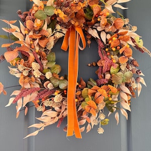 May include: A circular wreath featuring a mix of dried leaves in shades of orange, brown, and muted green, accented with small pinecones, berries, and feathery plumes. A long, solid orange velvet ribbon hangs from the top center of the wreath.