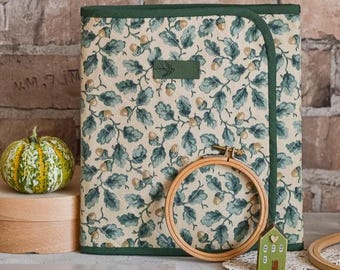 May include: A rectangular quilted fabric folder featuring a cream background with a repeating pattern of green oak leaves and brown acorns. The folder has dark green trim, a small rectangular label, a wooden embroidery hoop, and a small house charm.