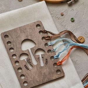 May include: A wooden embroidery floss organiser featuring a mushroom design, with brown, blue, and orange embroidery floss. The organiser rests on white fabric, with wooden embroidery hoops and buttons visible in the background.
