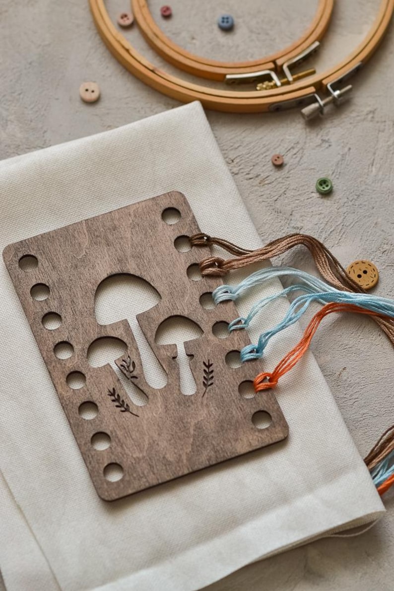 May include: A wooden embroidery floss organiser featuring a mushroom design, with brown, blue, and orange embroidery floss. The organiser rests on white fabric, with wooden embroidery hoops and buttons visible in the background.