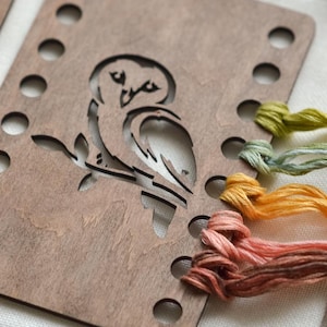 May include: A wooden embroidery hoop featuring an owl design. The brown wood has a series of holes for stitching. Various colours of embroidery thread, including orange, green, and pink, are also visible.