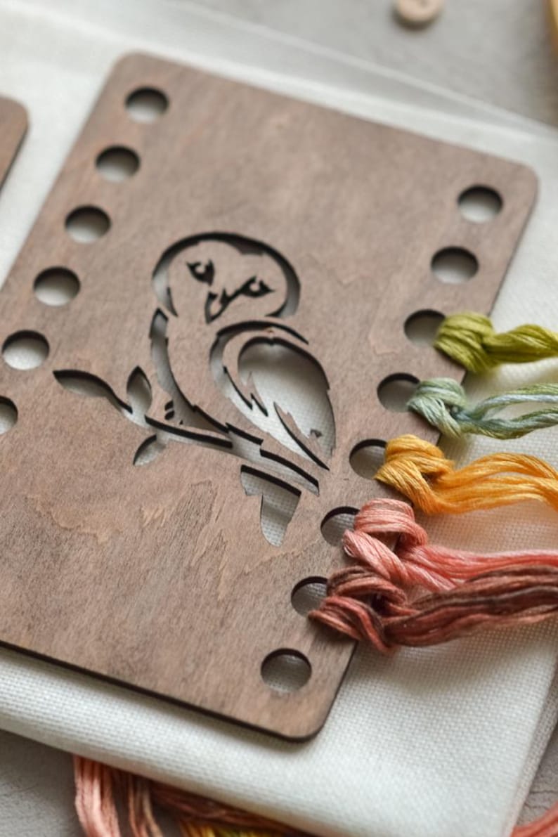 May include: A wooden embroidery hoop featuring an owl design. The brown wood has a series of holes for stitching. Various colours of embroidery thread, including orange, green, and pink, are also visible.