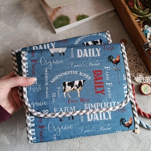May include: Two blue fabric pouches with a farm-themed print featuring cows, sheep, and roosters. The pouches have a gingham trim and the words "Daily," "Organic," and "Fresh" are printed on the fabric.