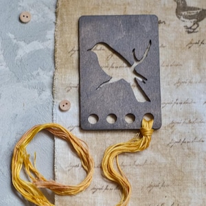 May include: A wooden bookmark with a bird silhouette, attached to yellow embroidery floss. The bookmark is gray and has three holes at the bottom. Two small wooden buttons and a bird illustration are also visible.