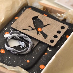 May include: A craft kit in a box, featuring a black and gray color scheme with a celestial theme. Includes a wooden bird-shaped embroidery floss holder, black quilted fabric, embroidery floss, and orange accents. The kit is designed for embroidery projects.