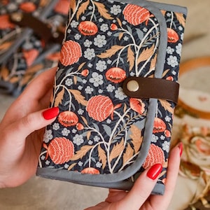 Könnte beinhalten: Eine Clutch mit floralem Muster auf dunkelblauem Hintergrund, mit orangefarbenen und weißen Blumen und hellbraunen Blättern. Die Clutch hat einen braunen Lederriemen mit silbernem Druckknopfverschluss und einen grauen Rand.