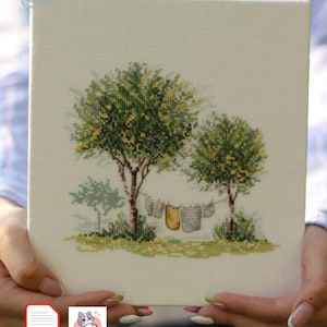 May include: Cross-stitch embroidery on canvas depicting a serene landscape with two trees, green foliage, and yellow accents. Clothes hang on a clothesline between the trees. The artwork is held by a person.