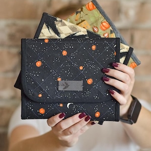 Peut inclure: Une pochette en tissu matelassé avec un fond noir, un motif d'étoiles blanches et des cercles orange. Le rabat avant présente une étiquette grise rectangulaire avec une icône d'oiseau. Une seconde pochette à motif de citrouilles est visible.