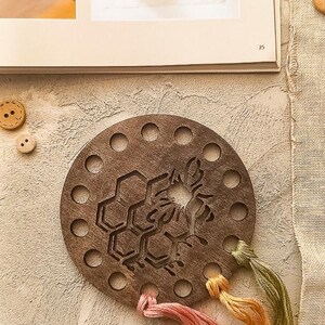 May include: A circular wooden embroidery floss organiser featuring a bee and honeycomb design. The organiser has multiple holes around the edge and is accompanied by strands of pink, yellow, and green embroidery floss. A book page is in the background.