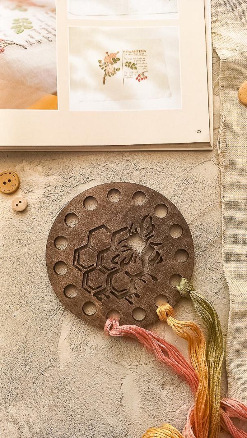 May include: A circular wooden embroidery floss organiser featuring a bee and honeycomb design. The organiser has multiple holes around the edge and is accompanied by strands of pink, yellow, and green embroidery floss. A book page is in the background.