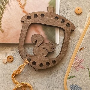 Peut inclure: Porte-fil à broder en forme de gland en bois avec une découpe d'écureuil. Le support est marron avec un petit trou pour le fil. Une écheveau de fil à broder doré est attaché. Deux boutons en bois et un projet de point de croix sont également visibles.
