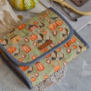 Puede incluir: Un estuche de tela acolchada con un estampado de temática otoñal con calabazas, pavos y granjas. El estuche tiene un borde gris y una etiqueta de cuero marrón. Una pequeña calabaza y un bastidor de bordado están en el fondo.