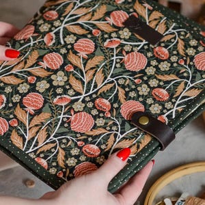 May include: A square, floral-patterned case with a dark green border and a brown leather strap. The fabric features a repeating design of orange and white flowers and leaves on a dark green background. The case is held by a person.