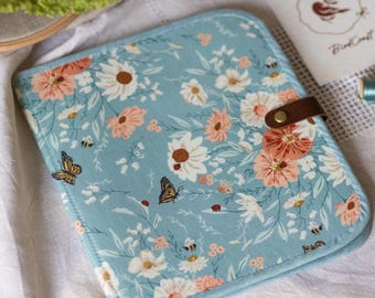 May include: A square fabric case in light blue featuring a floral pattern with white and peach flowers, butterflies, bees, and ladybugs. The case has rounded corners, a brown leather-like strap closure with a brass snap, and stitched edges.