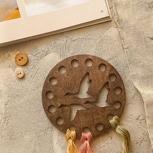 May include: A circular wooden thread organiser featuring a cut-out design of flying geese. It has multiple holes for holding thread spools and is paired with colourful embroidery floss in yellow, pink, and green. Two wooden buttons are also visible.