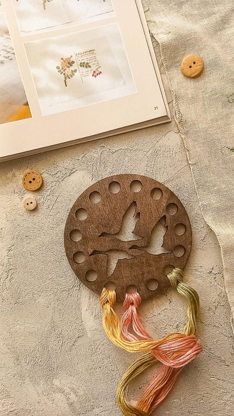 May include: A circular wooden thread organiser featuring a cut-out design of flying geese. It has multiple holes for holding thread spools and is paired with colourful embroidery floss in yellow, pink, and green. Two wooden buttons are also visible.