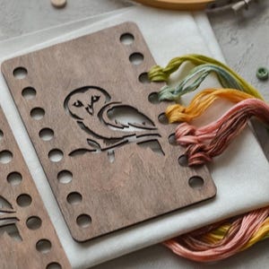 May include: A wooden embroidery hoop and a wooden frame with an owl cutout. Multicoloured embroidery threads in yellow, orange, and pink. Small wooden buttons and white fabric are also visible.