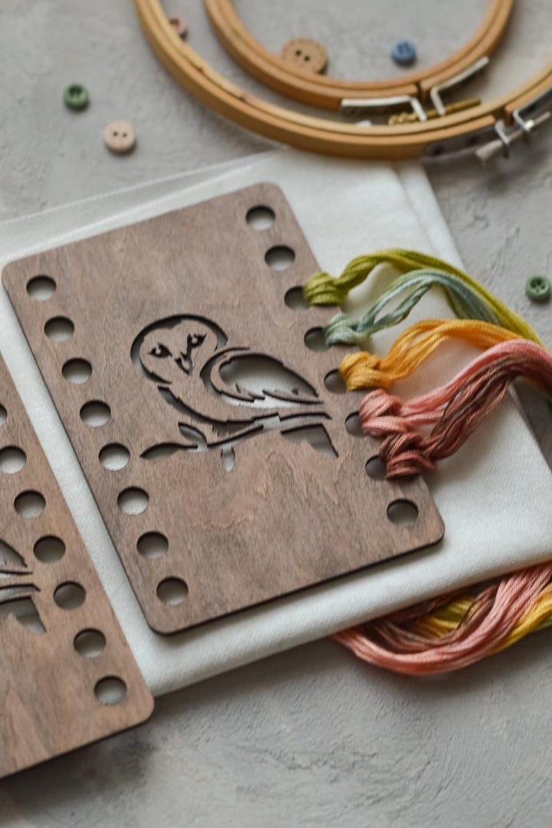 May include: A wooden embroidery hoop and a wooden frame with an owl cutout. Multicoloured embroidery threads in yellow, orange, and pink. Small wooden buttons and white fabric are also visible.