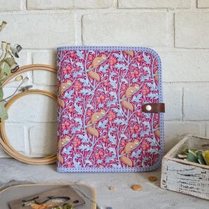 May include: A fabric-covered organizer with a colorful pattern of squirrels, flowers, and leaves in shades of pink, blue, and tan. The organizer has a brown leather strap closure and a light blue border.