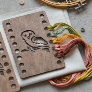 May include: A wooden embroidery hoop and an owl-shaped wooden template are on white fabric. There is colourful embroidery floss in yellow, orange, and pink, plus small buttons. The items are arranged on a grey surface, ideal for craft projects.