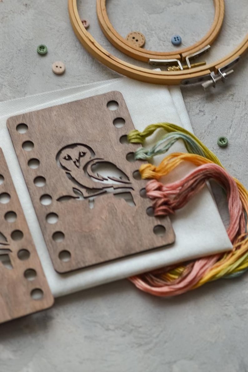 May include: A wooden embroidery hoop and an owl-shaped wooden template are on white fabric. There is colourful embroidery floss in yellow, orange, and pink, plus small buttons. The items are arranged on a grey surface, ideal for craft projects.