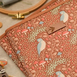 May include: Brown fabric with a repeating pattern of birds, mushrooms, and berries. The fabric is trimmed with a brown border. A small leather tag with a bird silhouette is attached. Embroidery hoop and scissors are in the background.