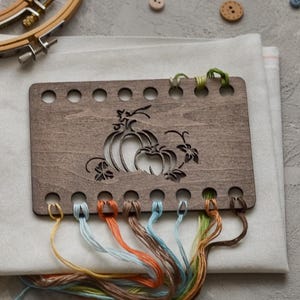 May include: A wooden cross-stitch holder with a pumpkin design, featuring various coloured threads. The holder is placed on white fabric, with embroidery hoops and buttons in the background. The threads are in shades of blue, orange, and brown.
