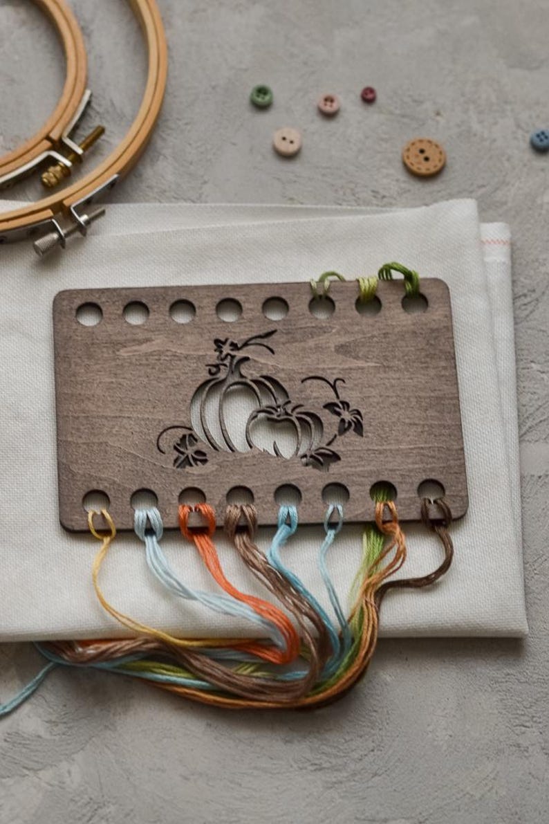 May include: A wooden cross-stitch holder with a pumpkin design, featuring various coloured threads. The holder is placed on white fabric, with embroidery hoops and buttons in the background. The threads are in shades of blue, orange, and brown.
